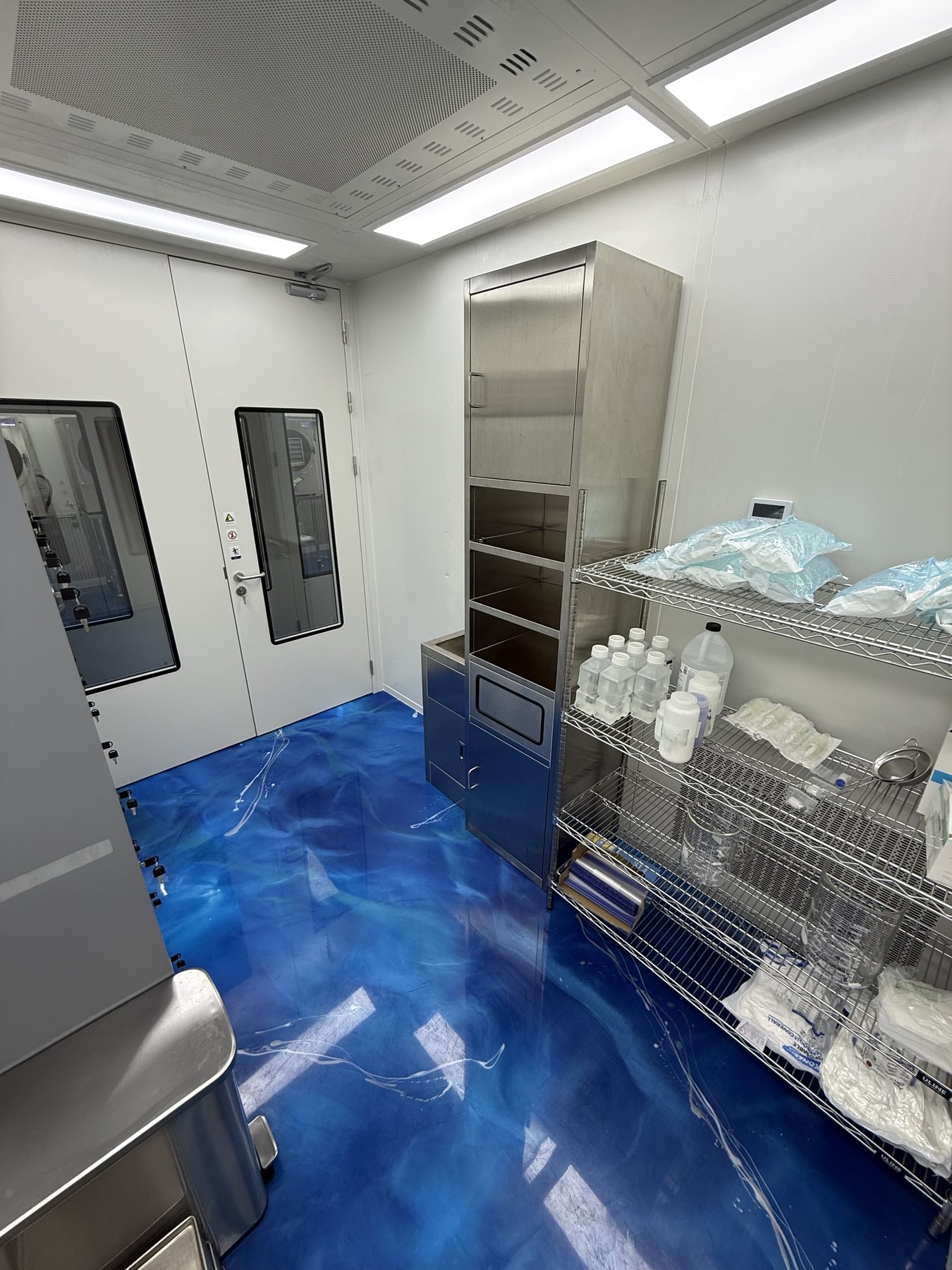 Alpha Peps cleanroom staging area with pre-sterilized pouched components, stoppers, seals and bottles on stainless wire shelving beside a stainless cabinet.