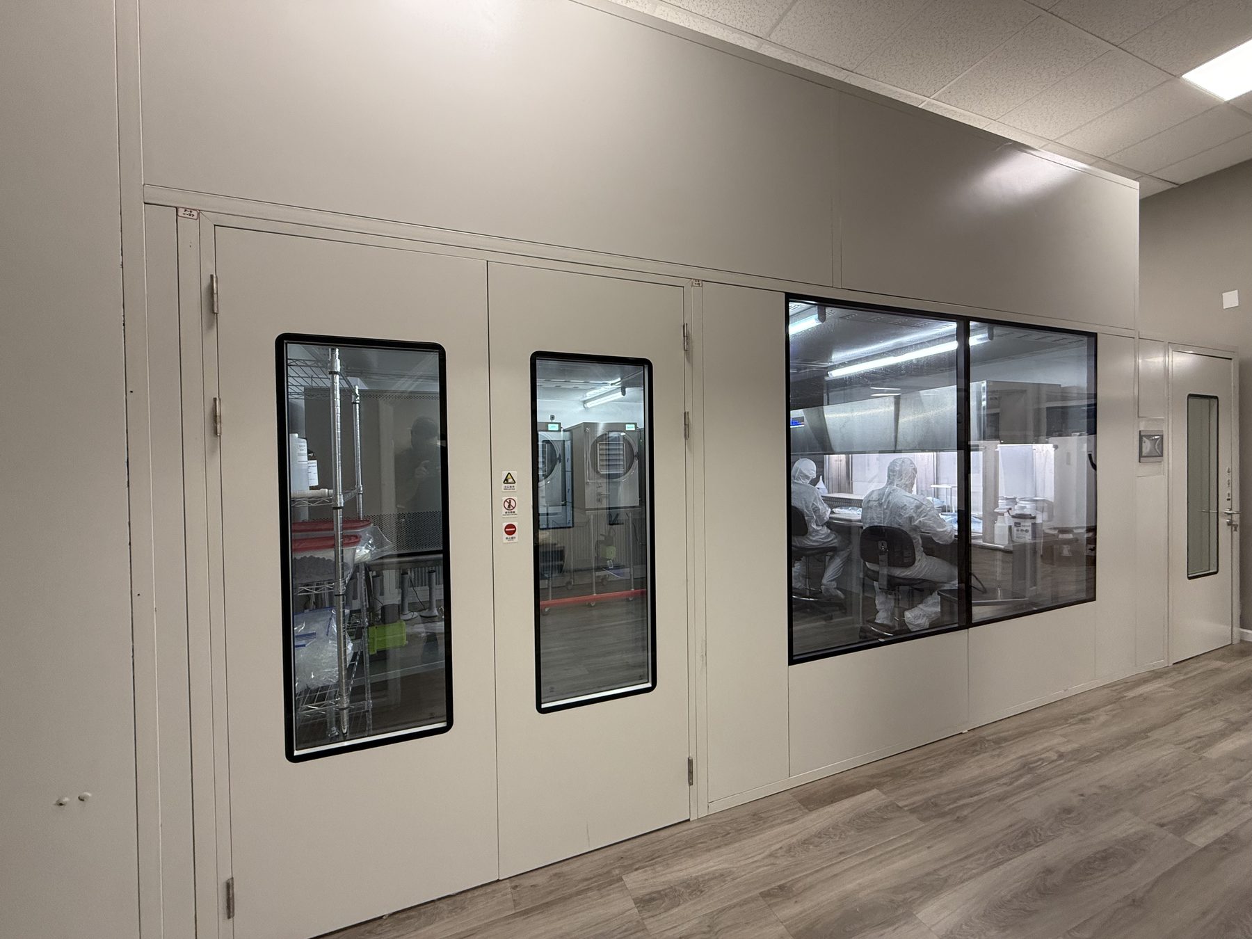 Alpha Peps modular hardwall cleanroom suite seen from the facility corridor, with sealed doors, viewing windows and gowned personnel working inside.