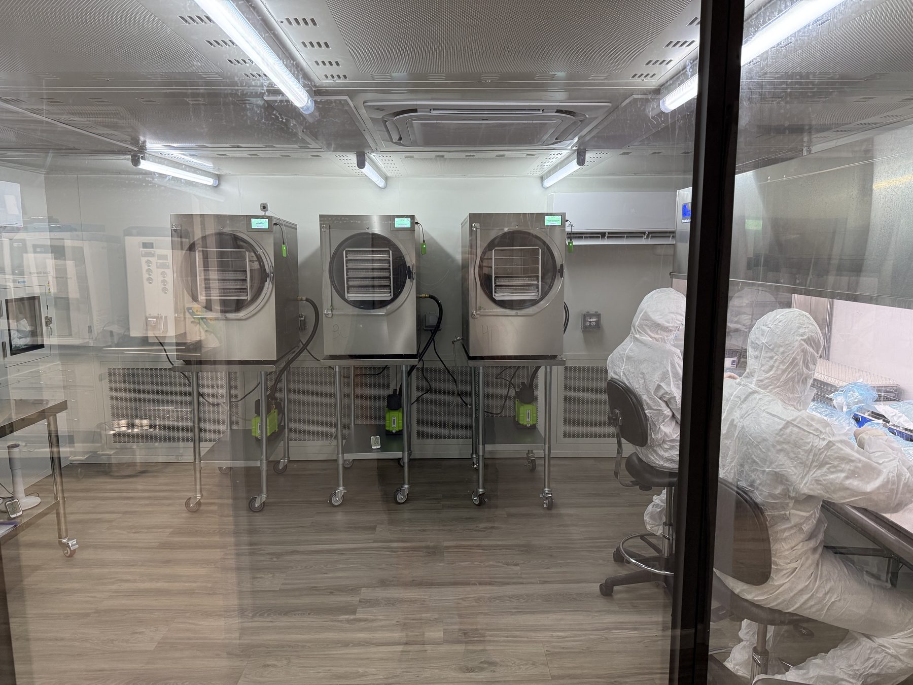 View through the cleanroom glass wall into the Alpha Peps lyophilization area, with three freeze dryers in the background and gowned operators working at a bench.